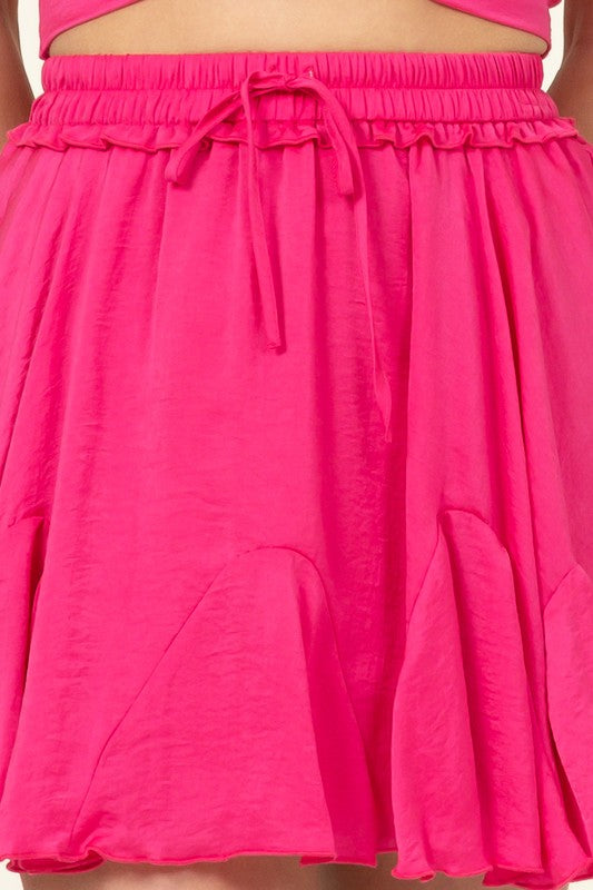 A person is partially visible in the image wearing the Not Your Girl Drawstring Ruffled Mini Skirt, featuring a bright pink flared silhouette with an elastic waistband and drawstring, paired with a matching top.