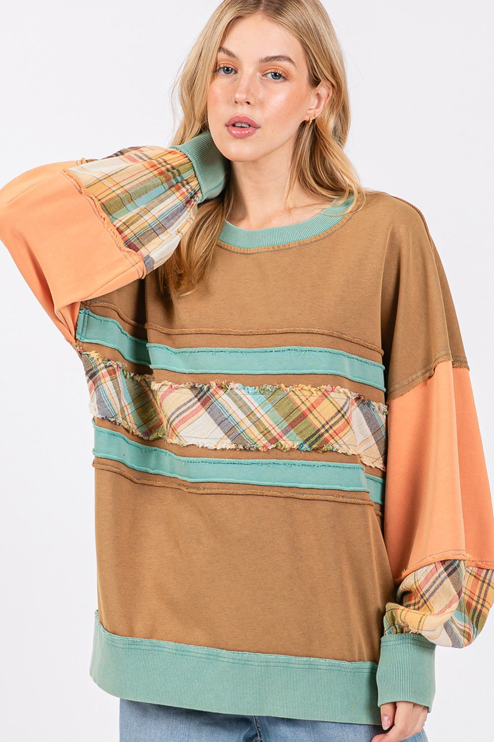 A person wearing a brown hat and the SAGE + FIG Mineral Wash Raw Edge Color Block Sweatshirt, featuring a patchwork design with plaid print contrast and solid color panels, smiles while posing with one hand in their pocket.