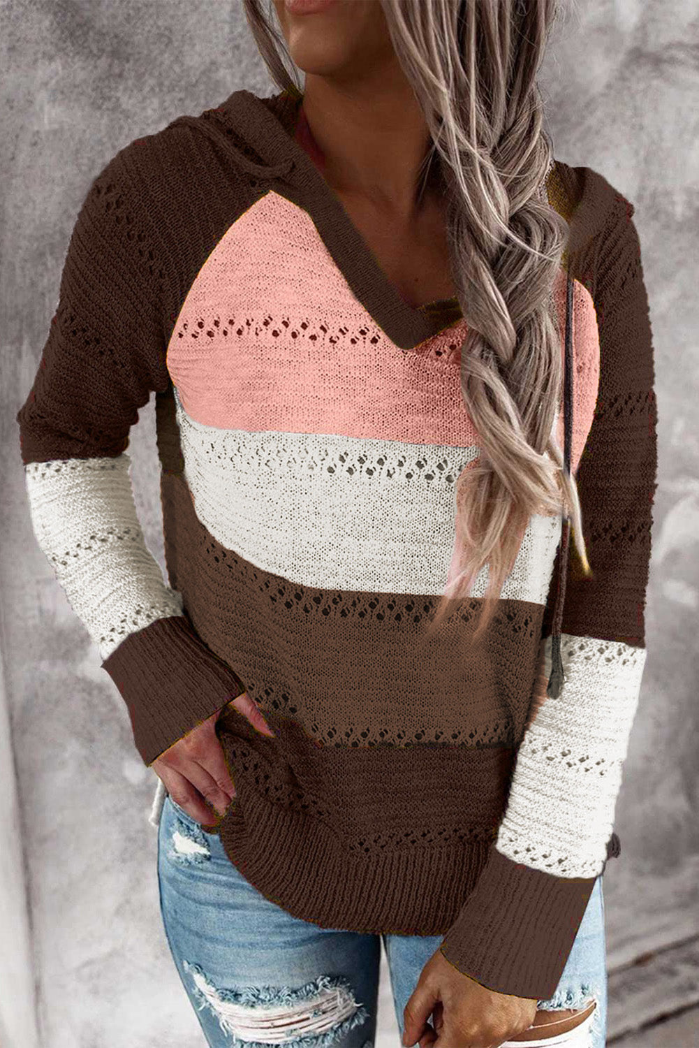 A person stands with their back to the camera, adjusting the hood of a Brown Beach Bonfire Knitted Hoodie featuring dark brown, pink, white, and light brown stripes. Perfect for a casual style, they are wearing distressed blue jeans and showcasing the laid-back vibe of lightweight hoodies.