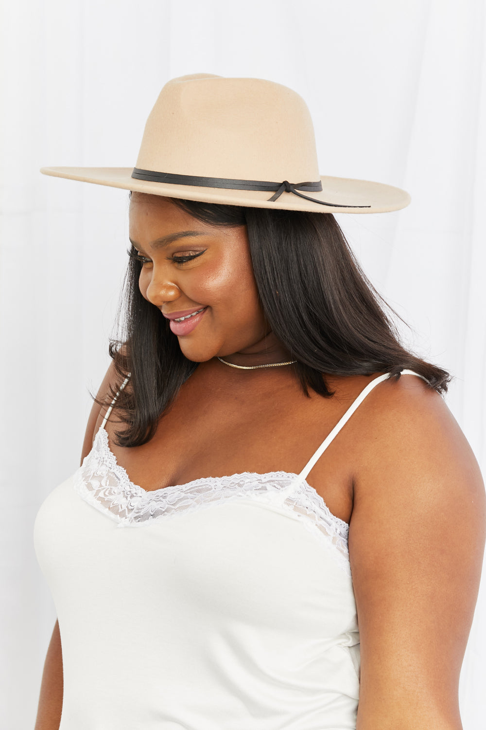 Wearing the Fame Make It Work Fedora Hat in light beige with faux leather detailing and a white tank top, a person smiles slightly while tilting their hat with one hand.