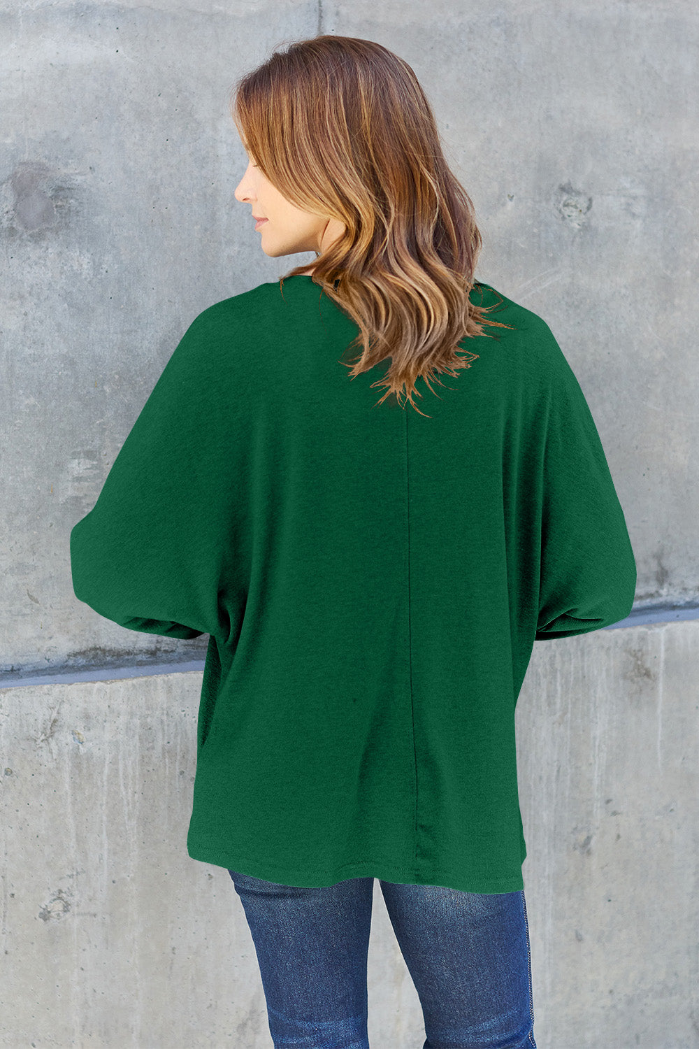 A person wearing a Double Take Full Size Round Neck Long Sleeve T-Shirt in green, blue distressed jeans with a slightly stretchy fit, and a crossbody bag, holding a beverage in a plastic cup.