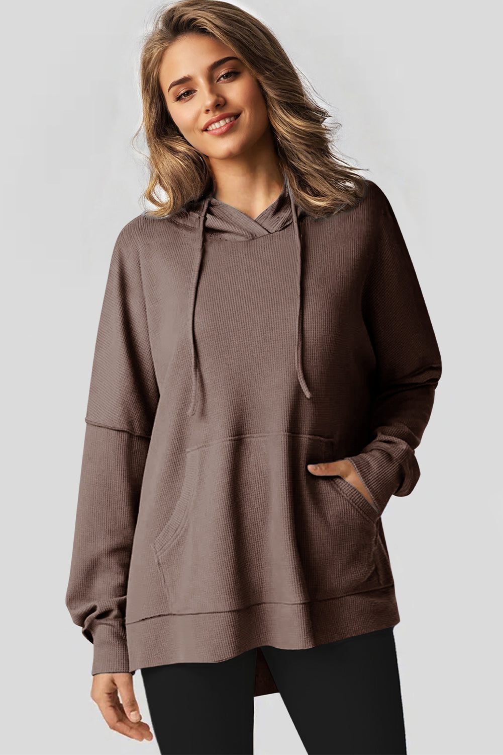 A woman with long hair is standing in profile, wearing a Coffee Waffle Knit Fleece Lined High Low Oversized Hoodie and black shorts. The background is a plain light gray.