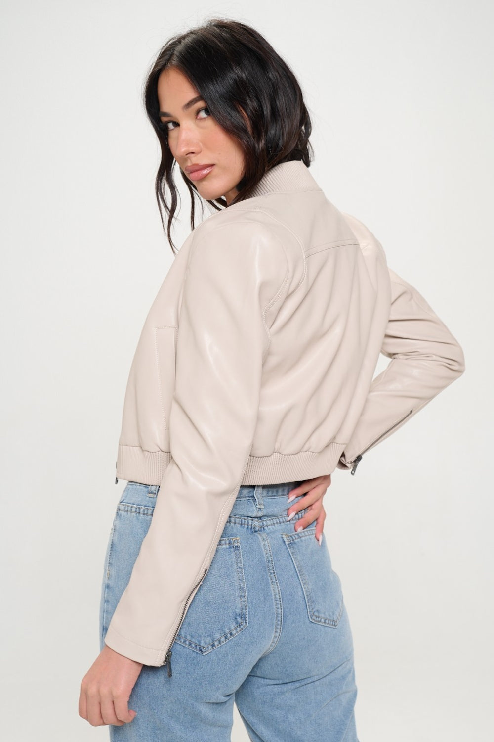 A woman wearing a Coalition LA Zip Up Cropped Bomber Jacket over a ribbed beige top and light blue jeans stands facing the camera, embodying sustainable fashion.