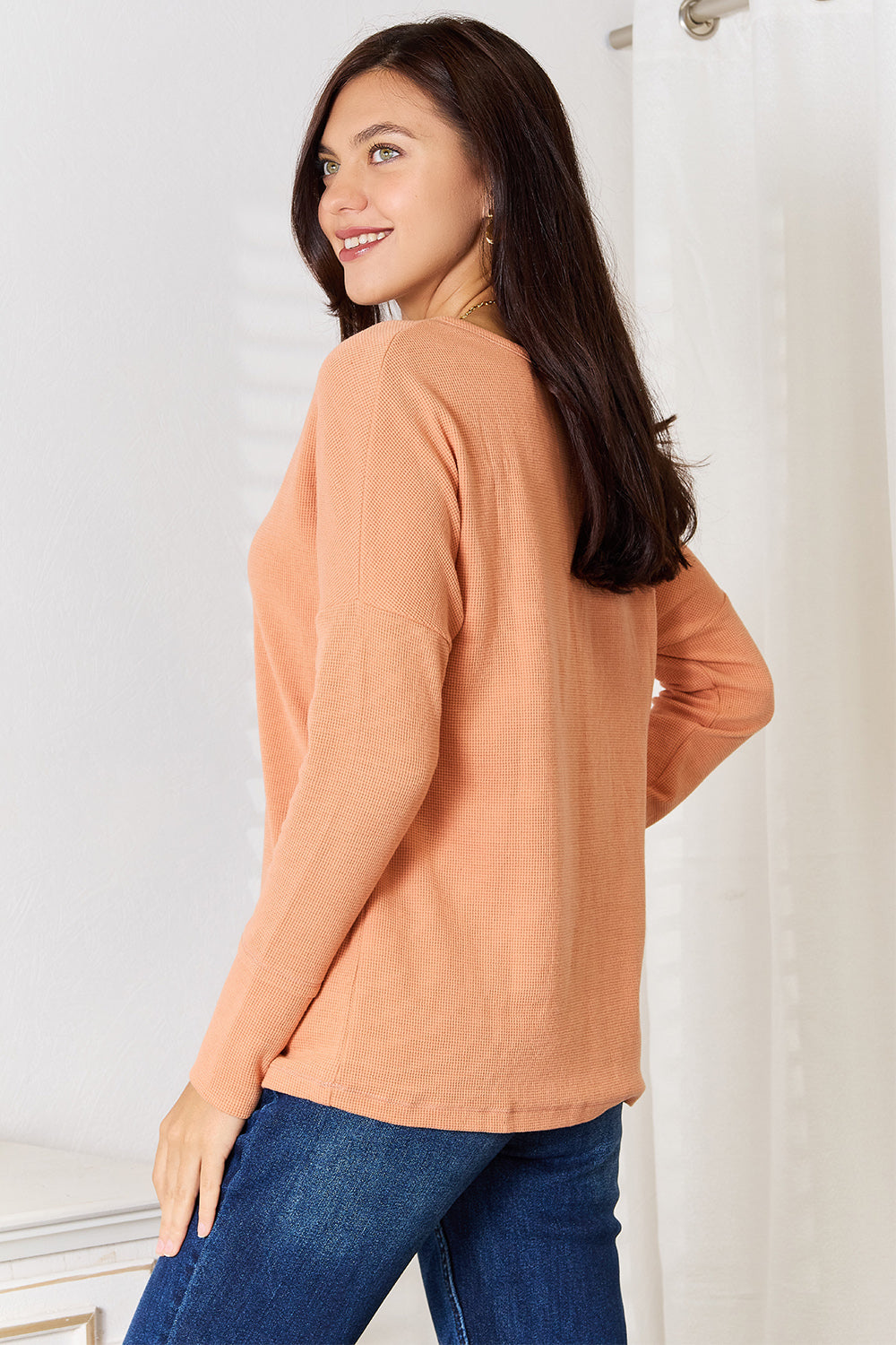 A woman stands indoors, wearing a Basic Bae Half Button Long Sleeve Top in peach and blue jeans, with her hand resting on her hip, showcasing versatile chic wardrobe choices.