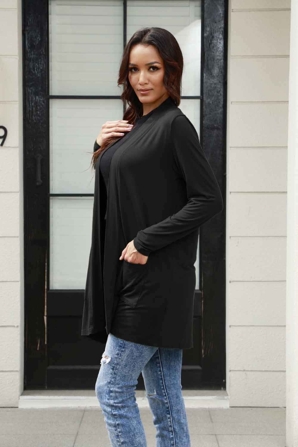 A person with long hair wears the Basic Bae Full Size Open Front Long Sleeve Cardigan with Pockets in gray, paired with a black shirt and blue jeans in a classic basic style. They stand in front of a building's glass door. The ensemble, made from 100% polyester, is perfect for those who prefer machine wash cold for easy care.