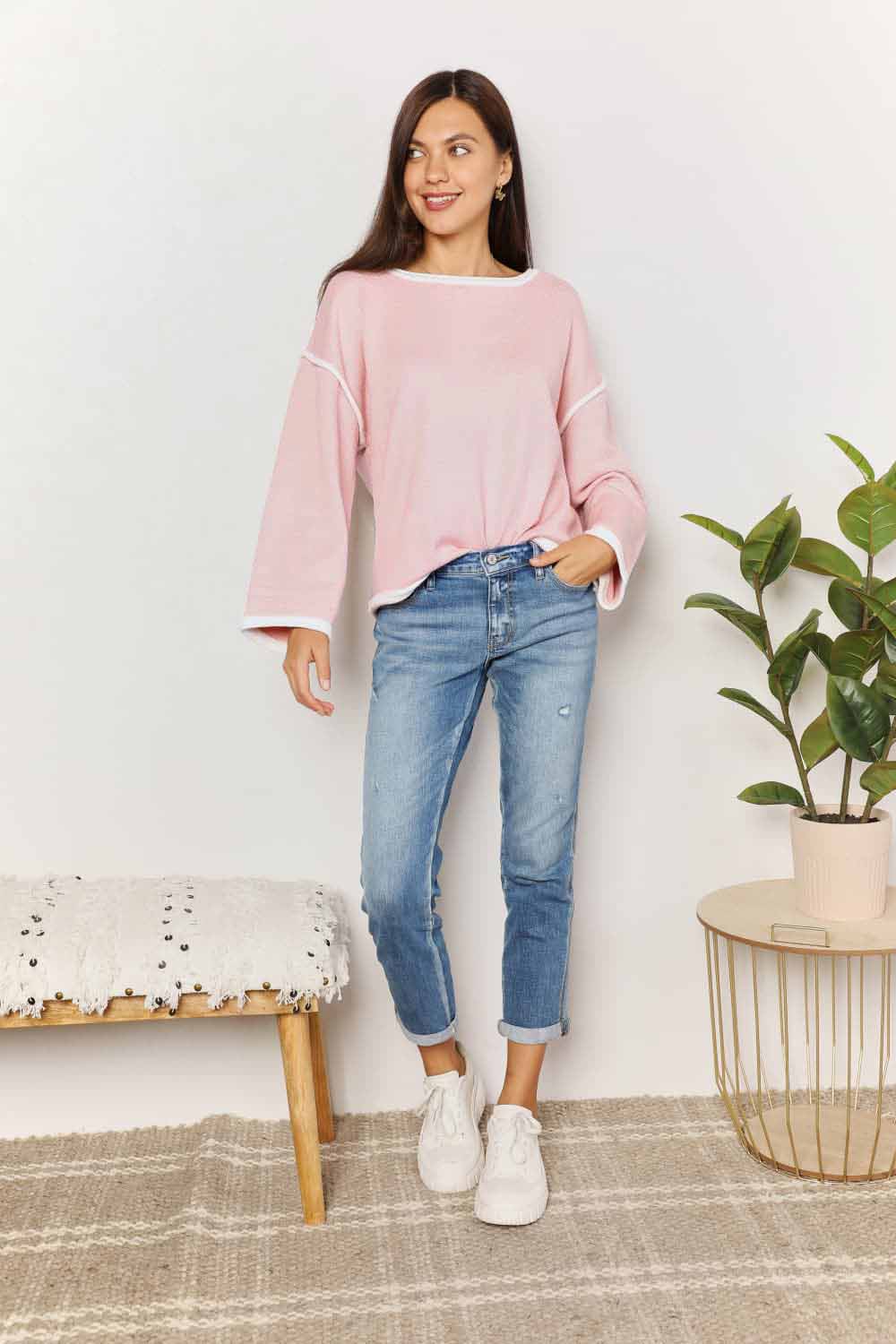 A woman with long curly hair is wearing the Angel Wings Contrast Detail Dropped Shoulder Knit Top in pink, paired with blue jeans, standing against a light background.