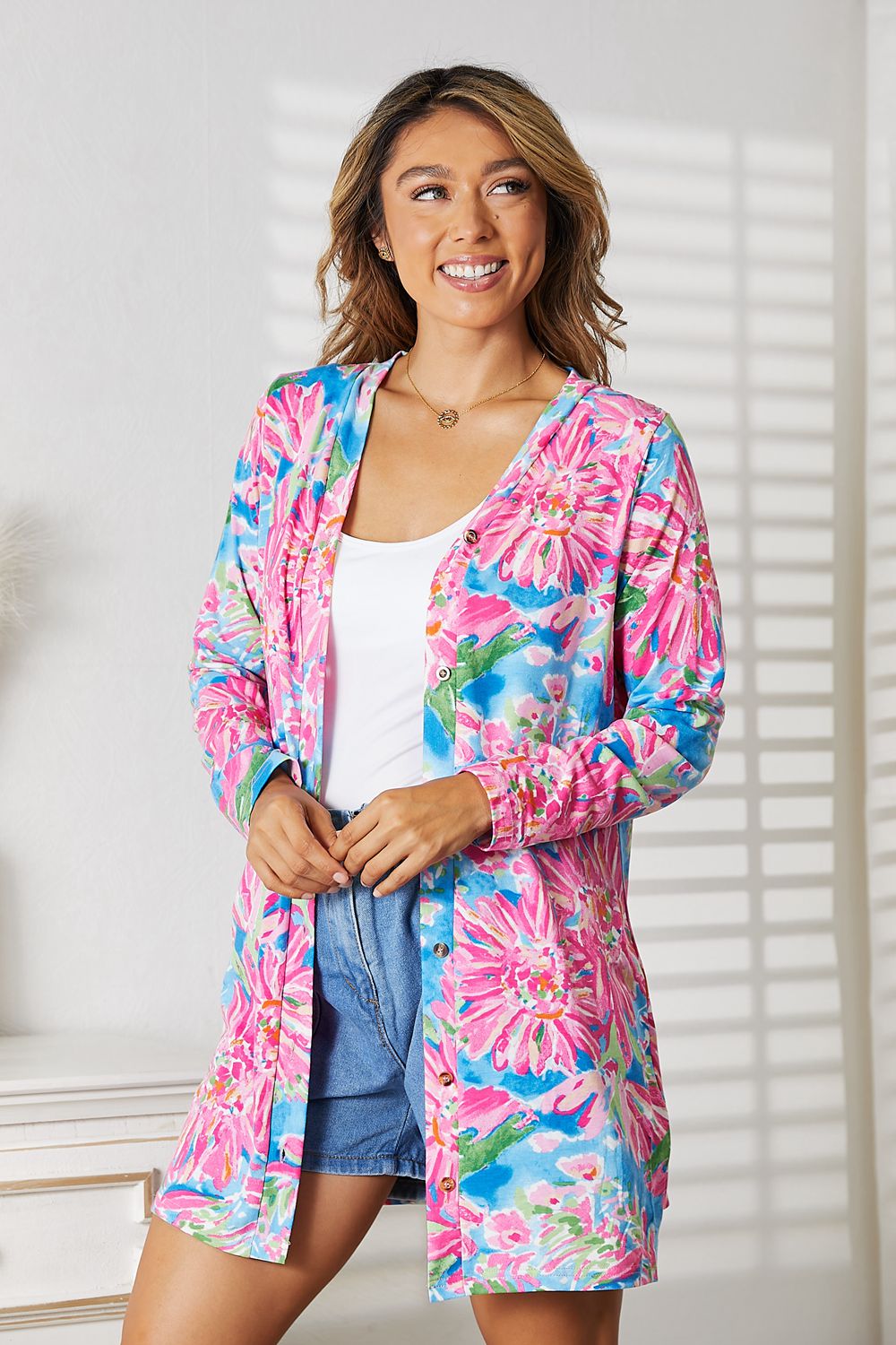 Wearing the Double Take Floral Open Front Long Sleeve Cardigan in shades of pink and blue over a white top and denim shorts, a person exudes feminine charm while standing in a well-lit room, smiling.