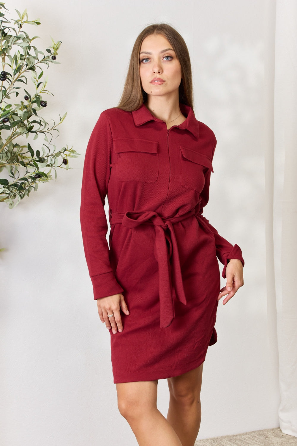 A woman wearing the Culture Code Full Size Tie Front Half Zip Long Sleeve Shirt Dress stands in front of a plain background next to a potted plant.