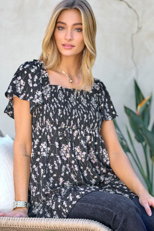 A woman stands outdoors donning a Floral Printed V-Neck Ruffle Top paired with jeans, set against a neutral background, with plants in the foreground, highlighting her fashion-forward wardrobe.