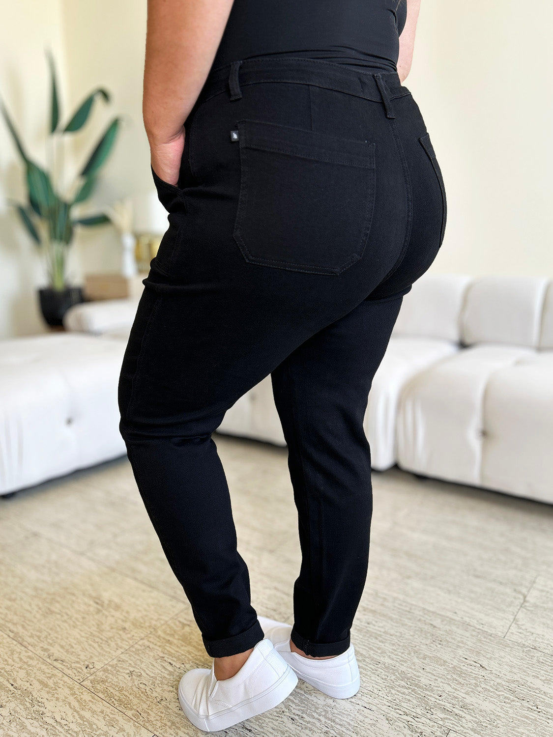 Person standing indoors, wearing a white top, black Judy Blue Full Size High Waist Double Roll Cuff Jeans, and white slip-on shoes, next to white couches and a plant in the background.