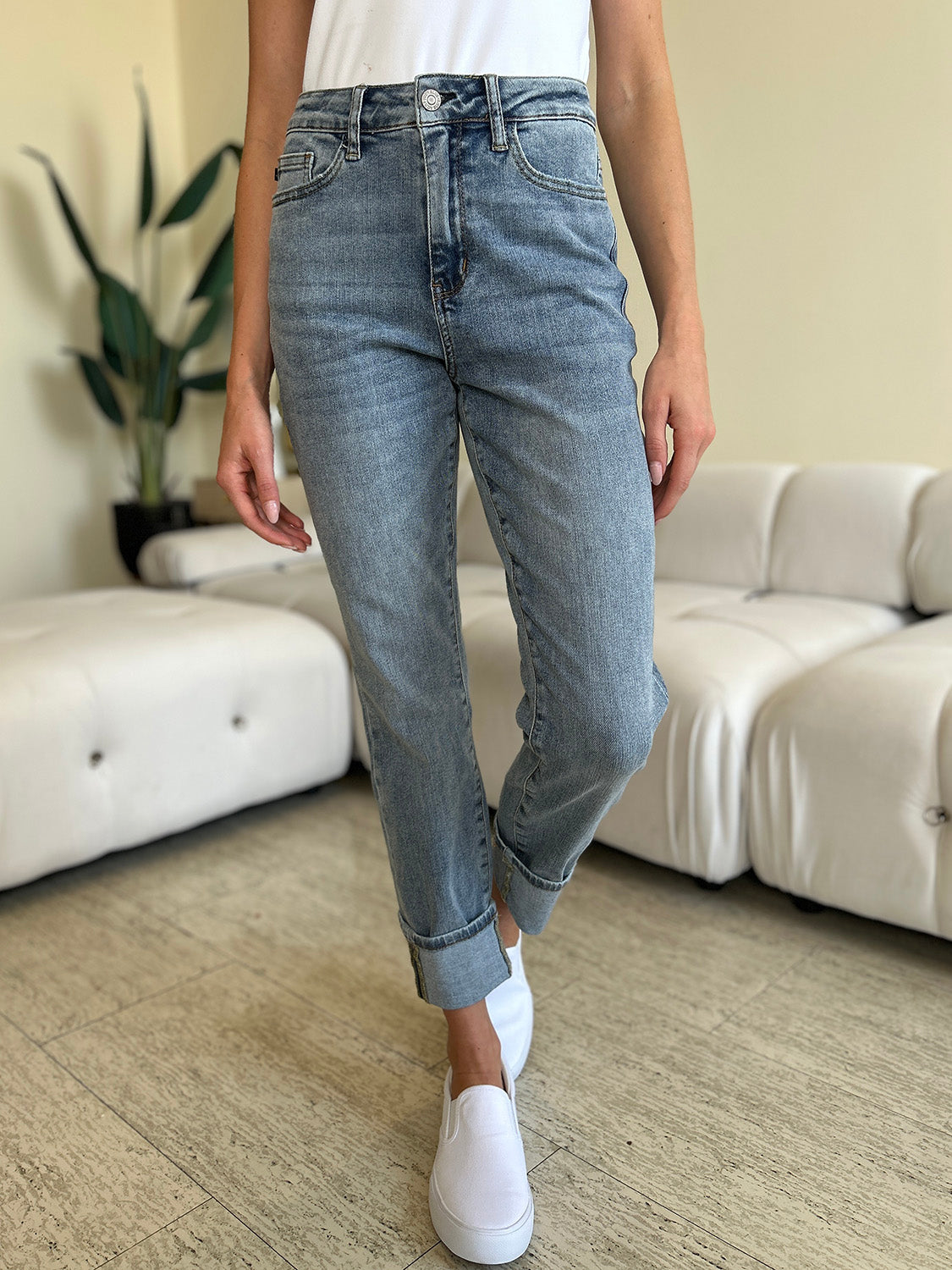 A person wearing Judy Blue Full Size High Waist Cuff Hem Jeans and white slip-on shoes stands in a living room with a white couch and plants in the background.