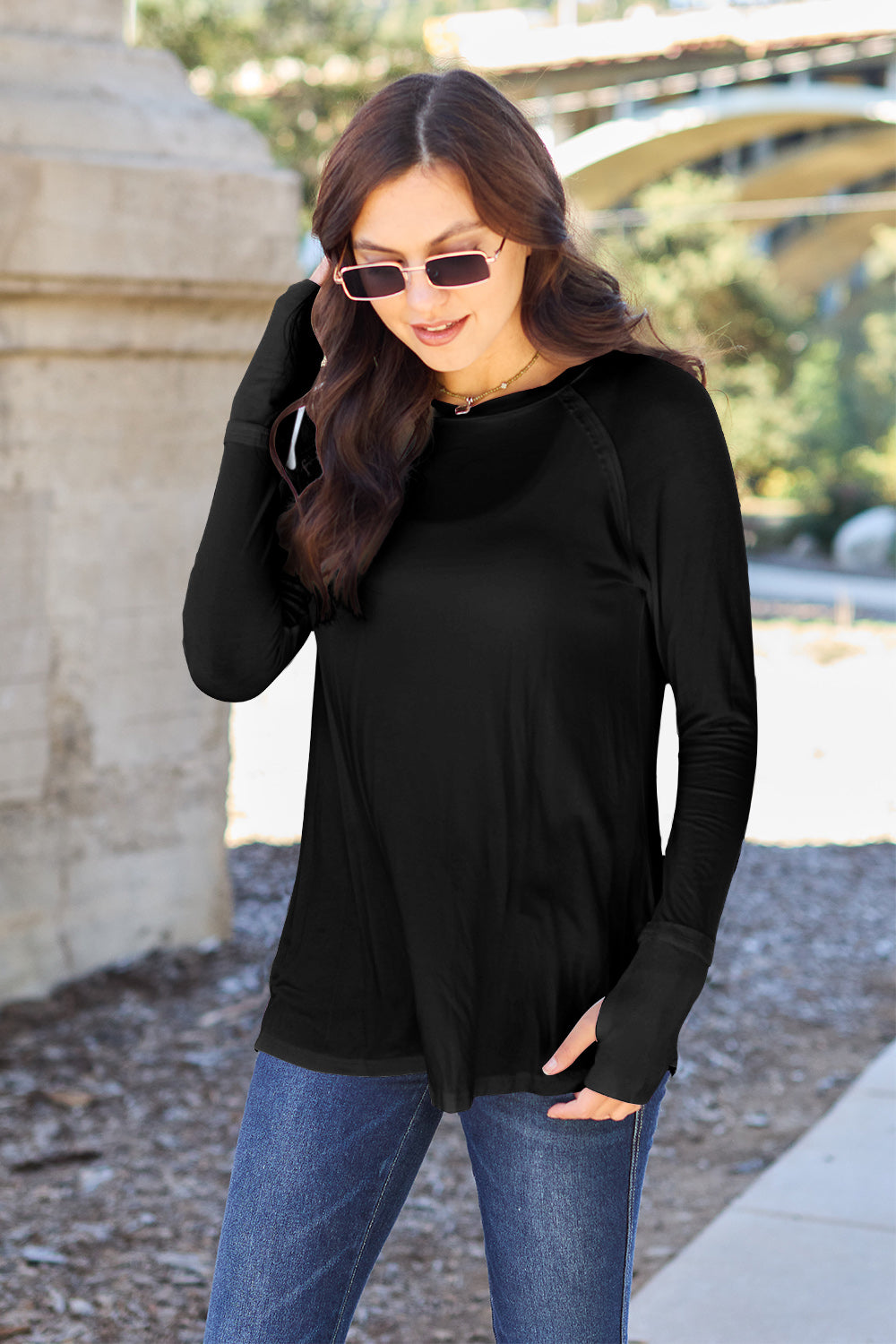 A woman wearing sunglasses and a slightly stretchy Basic Bae Full Size Round Neck Long Sleeve T-Shirt in white stands on a sidewalk with an arched structure in the background, smiling slightly.