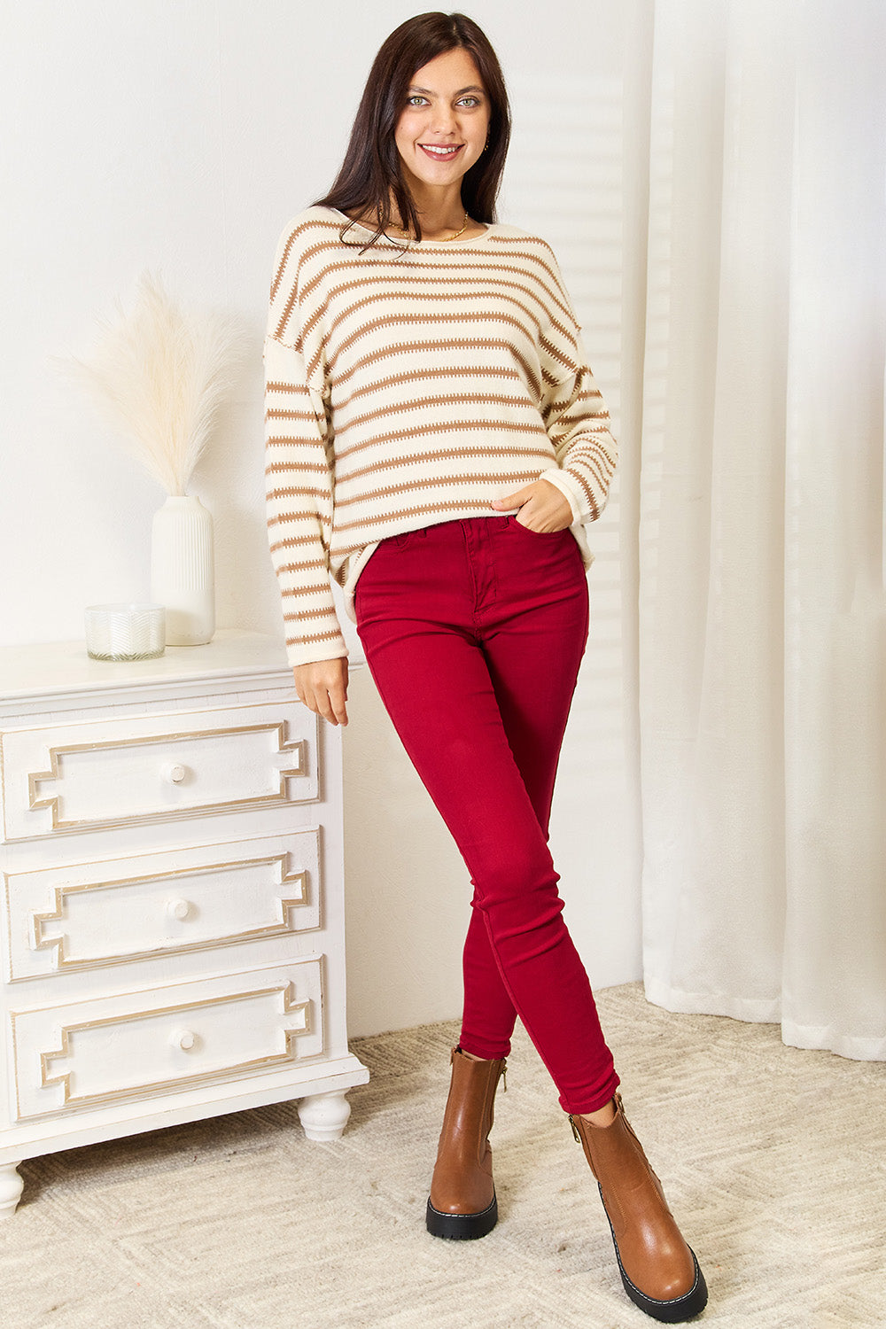 A woman stands indoors, smiling and looking to the side while wearing the Double Take Striped Boat Neck Sweater in cream and brown, paired with red pants.