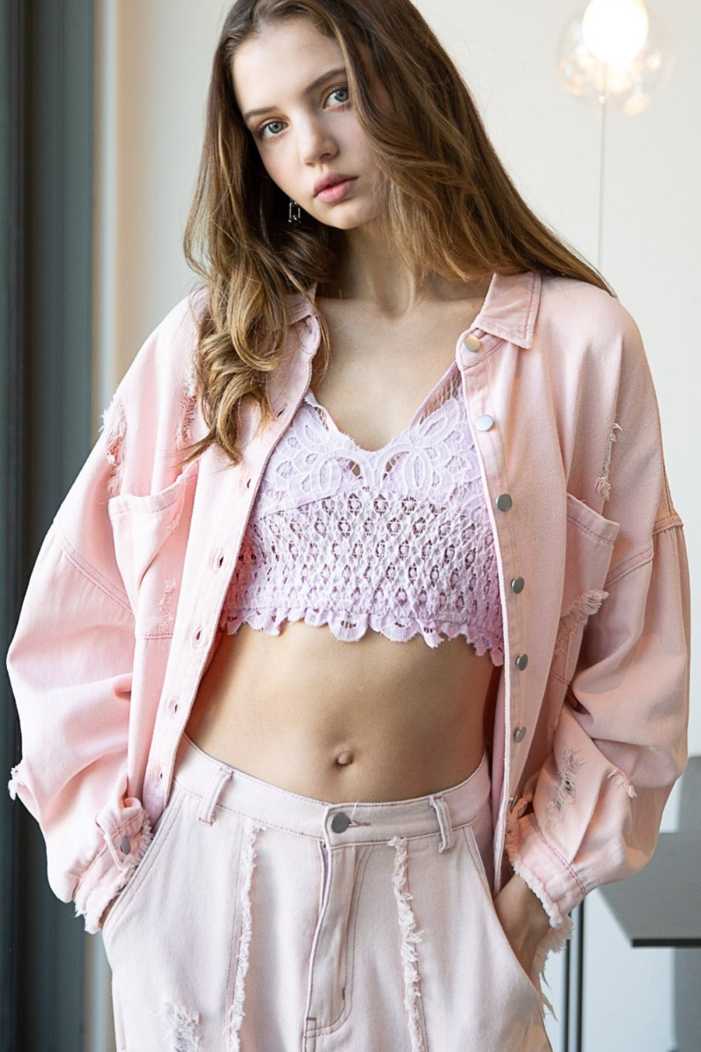 A woman wearing a POL Button Down Raw Hem Distressed Shacket in pale pink over a lavender lace crop top and matching pale pink distressed pants, standing indoors near a window.