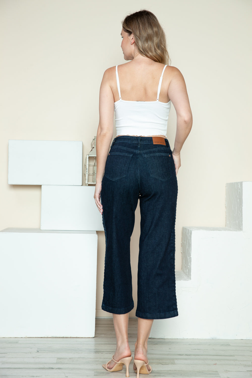 Person wearing Judy Blue Full Size Side Seam Braid Detail Crop Wide Leg Jeans and beige strappy heels, standing on a wooden floor with a white background.