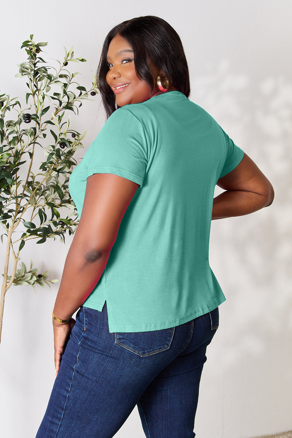 A person with wavy hair, wearing the Basic Bae Full Size Round Neck Short Sleeve T-Shirt in bright pink and beige pants, stands near a plant, smiling and looking to the side. The outfit embodies a basic style and offers slightly stretchy comfort, ideal for those who value effortless elegance.