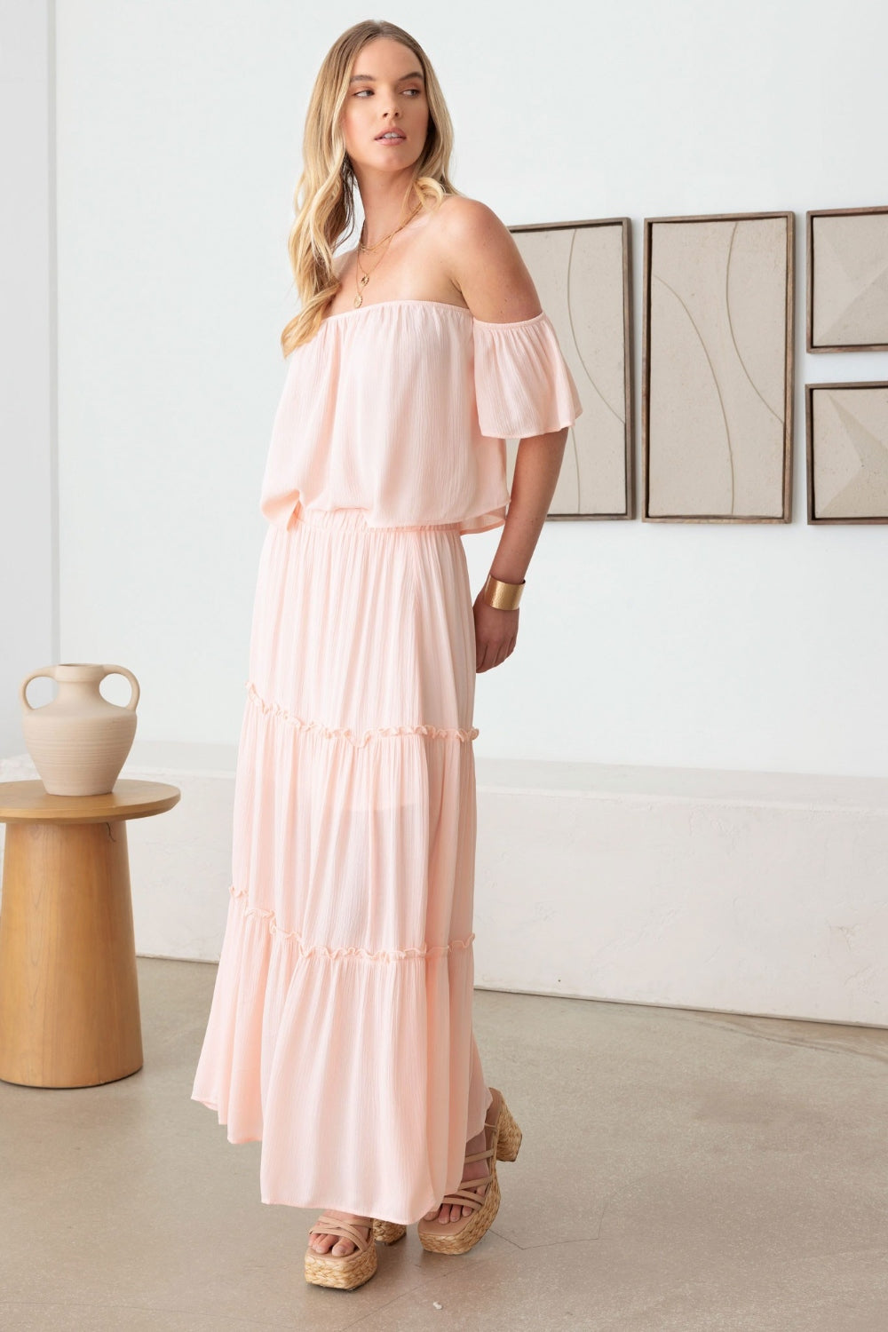 A woman in a romantic Gilli Frill Off-Shoulder Tiered Dress stands holding a beige woven handbag, with two abstract paintings on the wall behind her. She is wearing platform sandals.