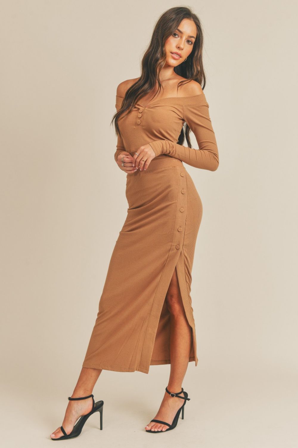 A woman stands against a plain background wearing the MABLE Button Detail Crop Top and Slit Midi Skirt Set, showcasing a camel-colored ensemble with off-shoulder, button details, and a side slit. She completes her look with black high-heeled sandals.