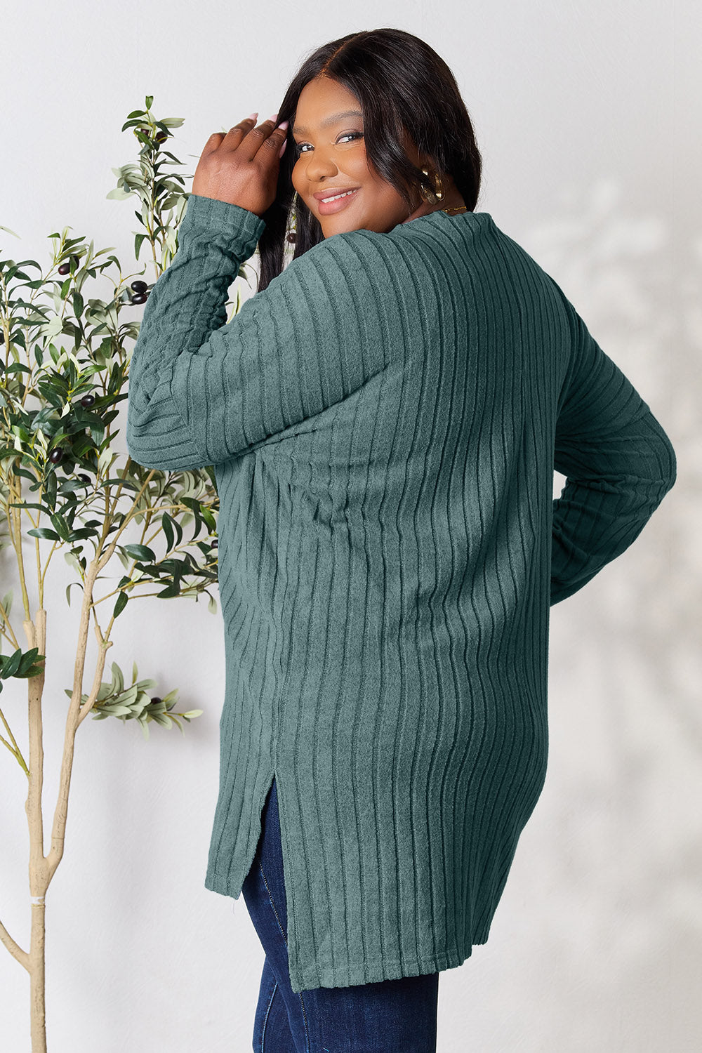 A person with shoulder-length hair smiles while wearing a beige ribbed sweater called the Basic Bae Full Size Ribbed Round Neck Long Sleeve Slit Top, along with matching beige pants, showcasing a basic style. They stand next to a green plant.