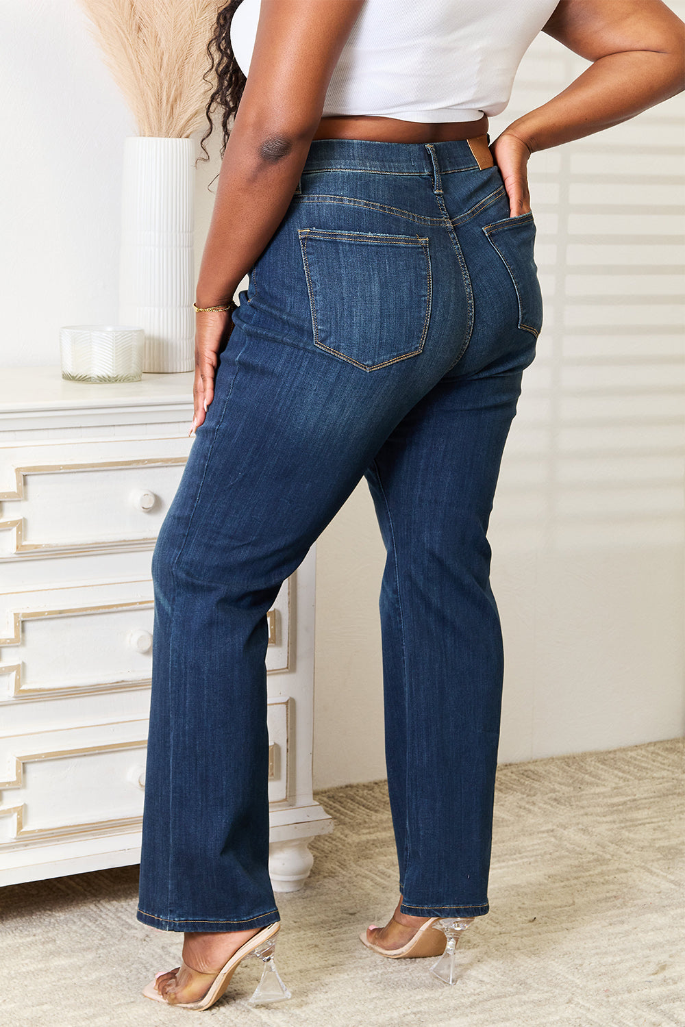 A person wearing a white crop top and Judy Blue Full Size Elastic Waistband Slim Bootcut Jeans stands next to a white dresser.