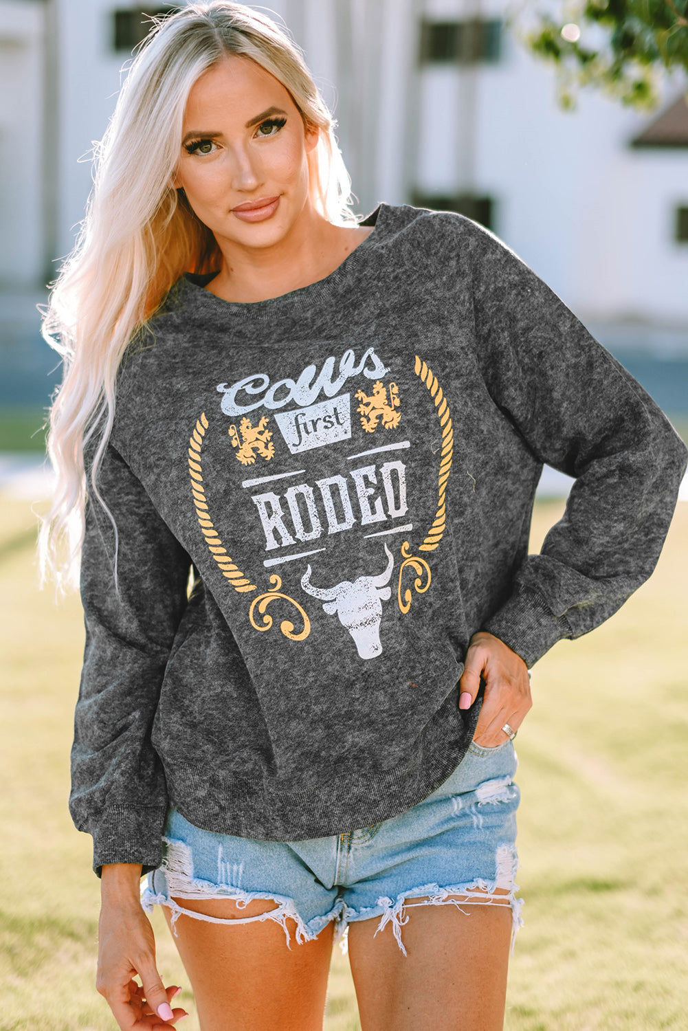 A blonde woman wearing a Gray Coors Banquet RODEO Graphic Mineral Washed Sweatshirt and light blue denim shorts, showcasing casual outdoor wear, stands outdoors with her back to the camera.