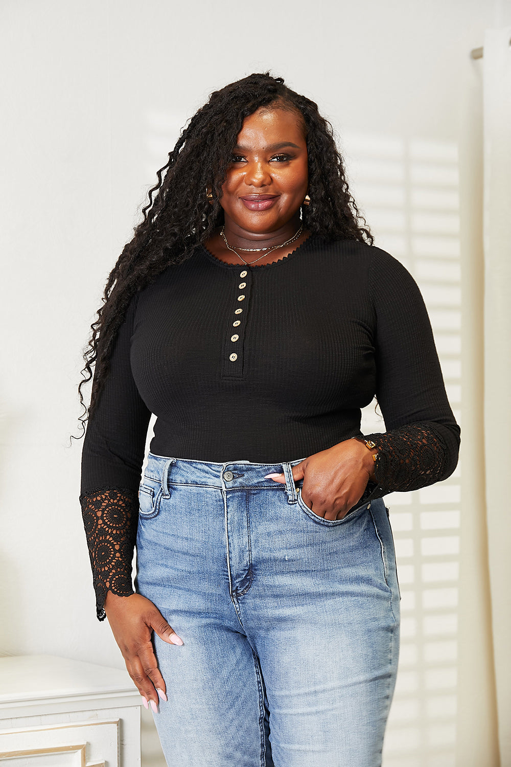 A woman with long curly hair wears the Double Take Crochet Lace Hem Sleeve Button Top and versatile solid color blue jeans, standing indoors with a neutral expression.