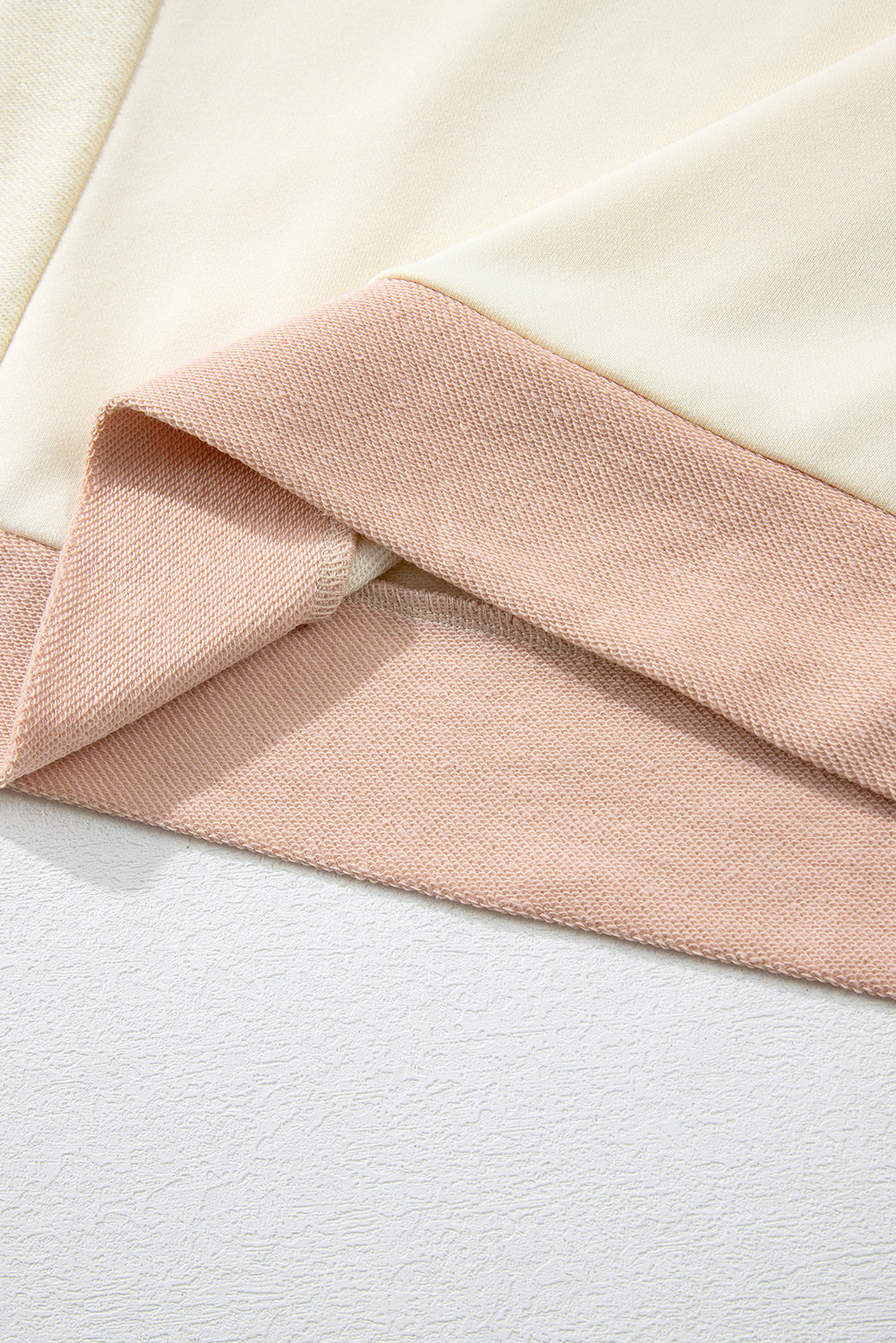 A person with long, wavy, light brown hair stands facing away, wearing the Beige Star Patchwork Exposed Seam Oversized Sweatshirt paired with denim shorts. The background is a plain, light peach wall, adding an edgy vibe to the look.