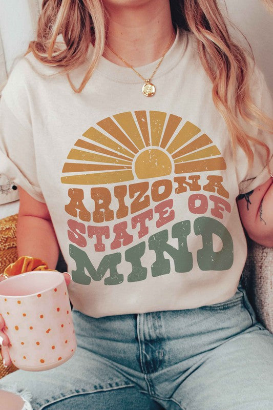 Person wearing the ARIZONA STATE OF MIND Graphic Tee, a beige 100% cotton t-shirt with "ARIZONA STATE OF MIND" text in brown and green colors. The unisex shirt offers both comfort and style for any occasion. They are holding a pink polka-dotted mug.