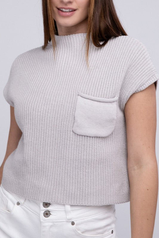 A woman in a JustFab mock neck short sleeve cropped sweater and white pants, smiling slightly, stands with one hand on her hip against a light grey background.