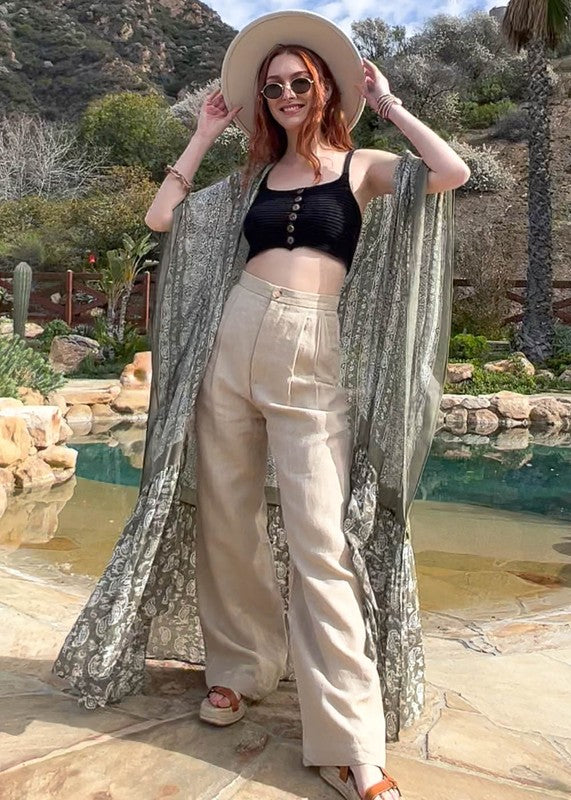 A woman wearing a Paisley Tapestry Free Flow Kimono stands in a bright, eclectic room with plants, books, and an oversized deer head on the wall. The space features large windows and industrial decor.