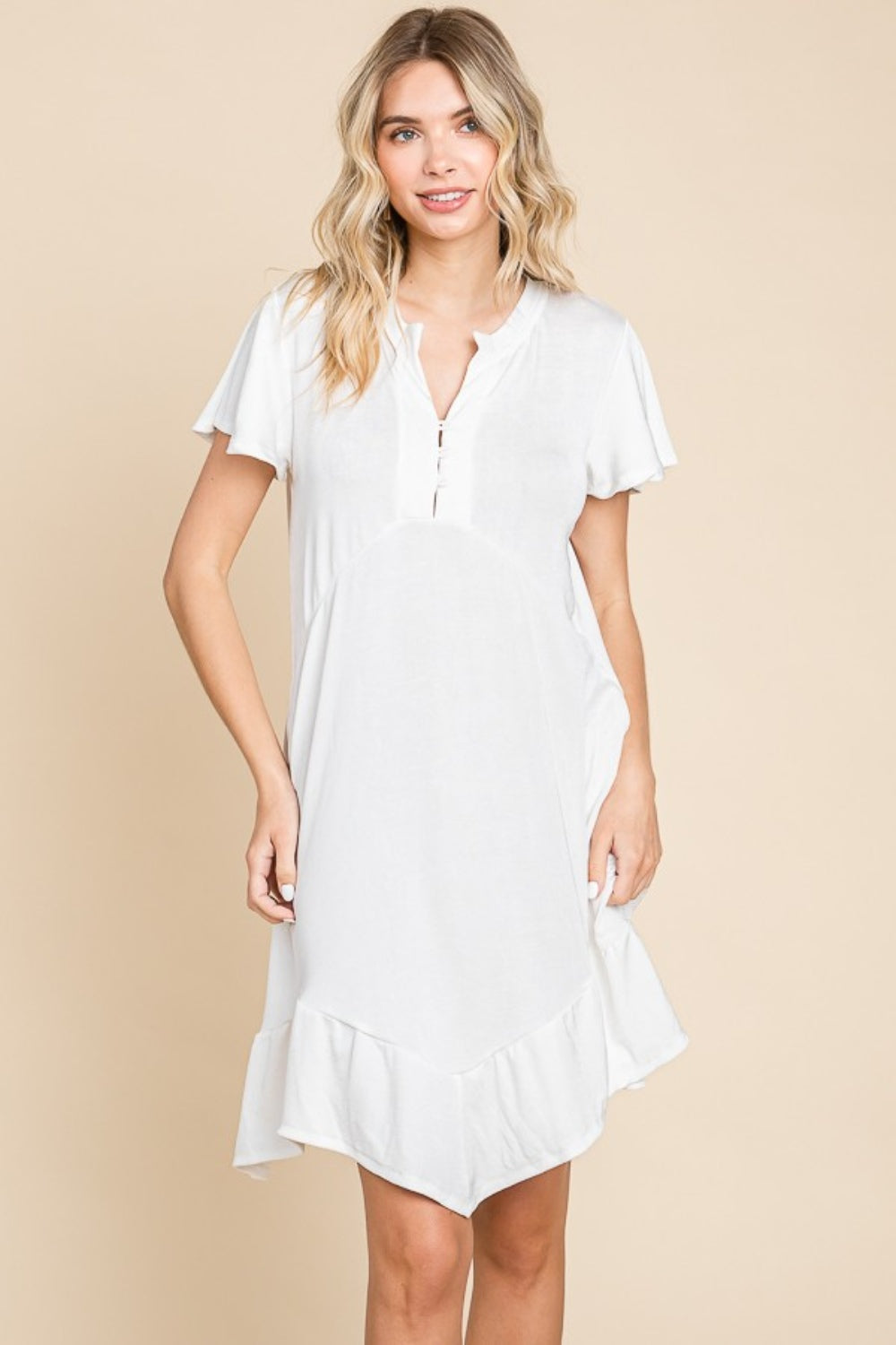 A person wearing the versatile white Culture Code Full Size Short Sleeve Ruffled Asymmetric Hem Dress stands in front of a plain beige background, showcasing its flattering fit.