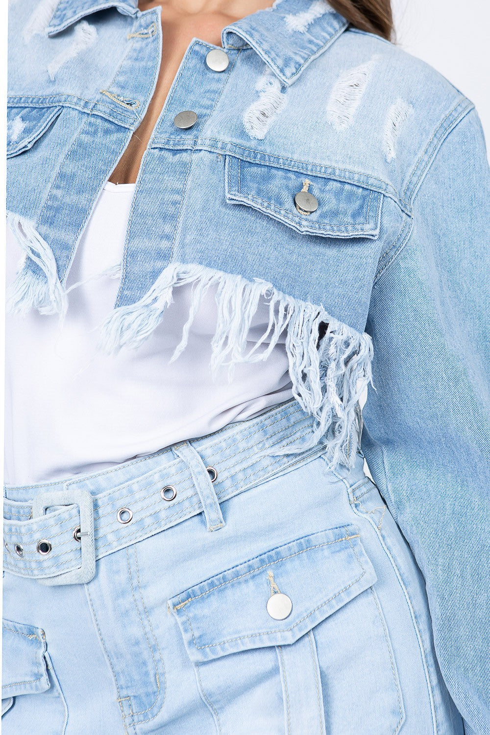 A woman with long brown hair wears an American Bazi Distressed Denim Jacket with Frayed Hem and a light denim skirt with front pockets. The jacket features frayed hems, adding a touch of edgy style. She stands facing the camera against a plain white background.