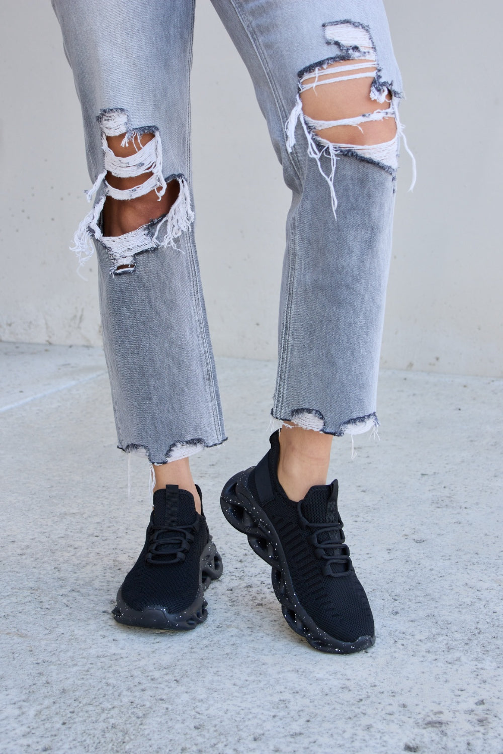 A person wearing ripped gray jeans and Forever Link Mesh Breathable Lace-Up Athletic Shoes is standing on a concrete surface.