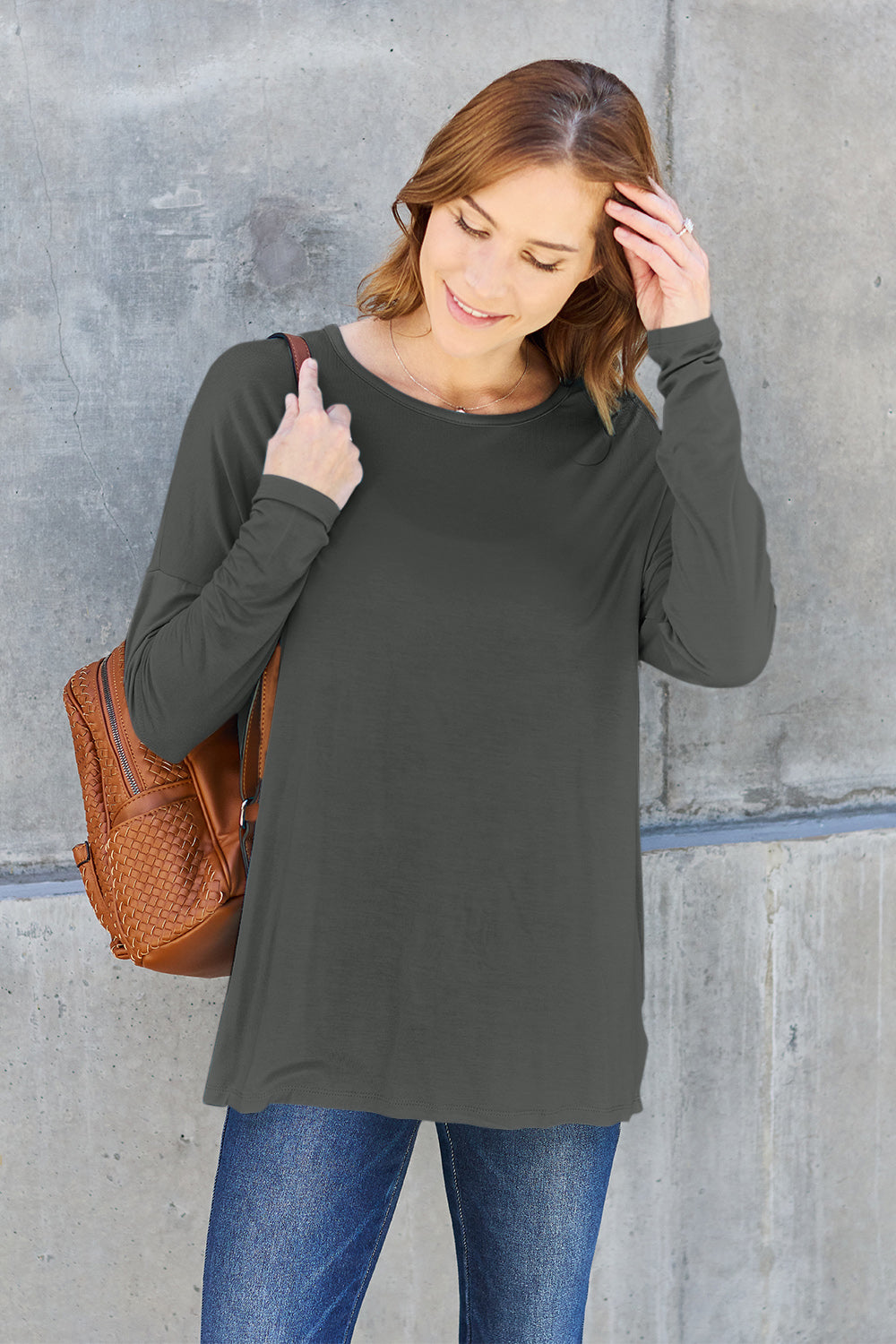 A woman with shoulder-length hair wearing a Basic Bae Full Size Round Neck Dropped Shoulder T-Shirt in green and a pair of jeans, exuding a basic style, stands against a concrete wall. She's holding an imported brown woven backpack on her shoulder while touching her hair with her hand.