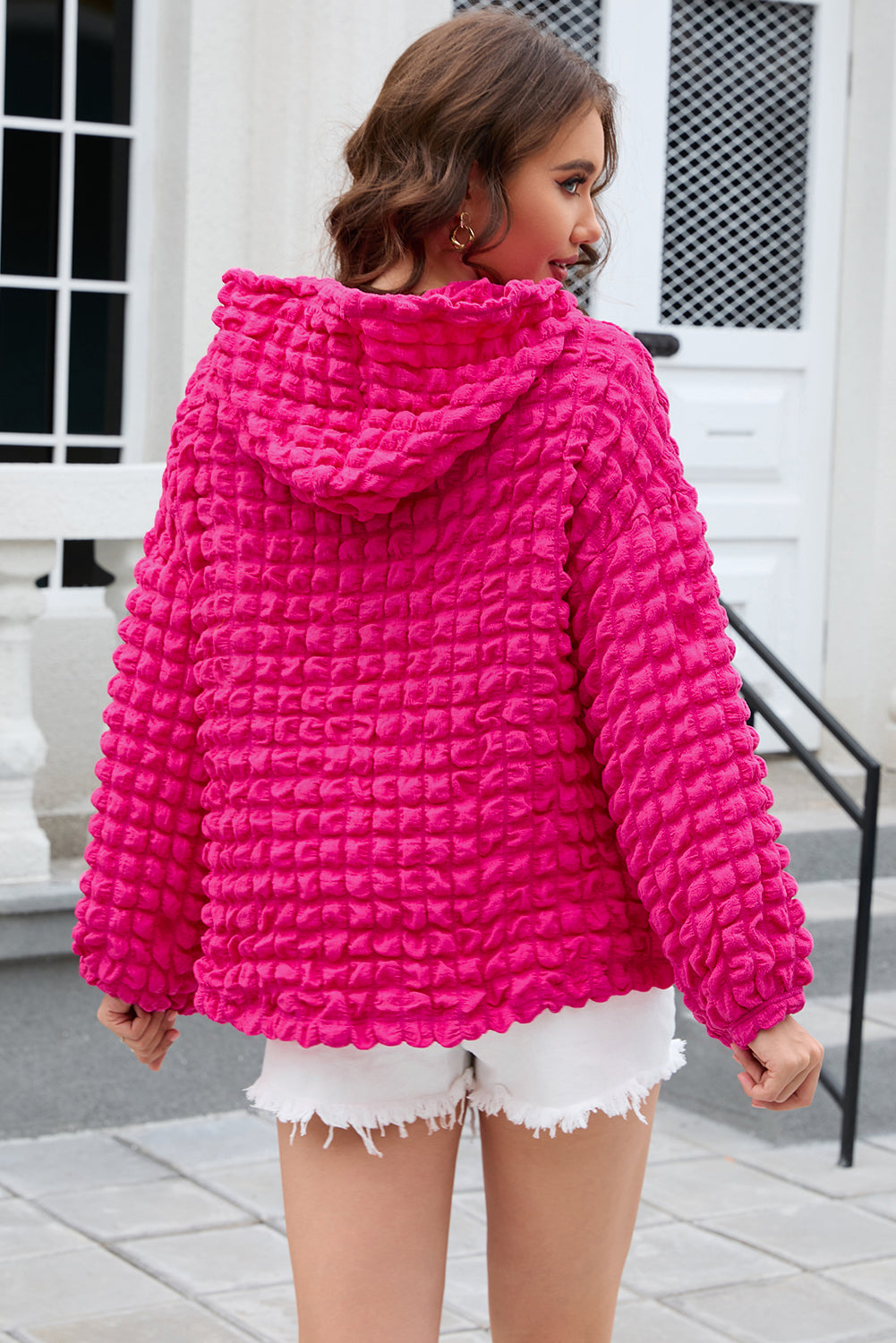 A person stands with their back to the camera, donning the Rose Bubble Textured Waffle Hoodie in a vibrant pink hue. They are in a room with yellow floral wallpaper, and a corner of a wooden chair is visible.