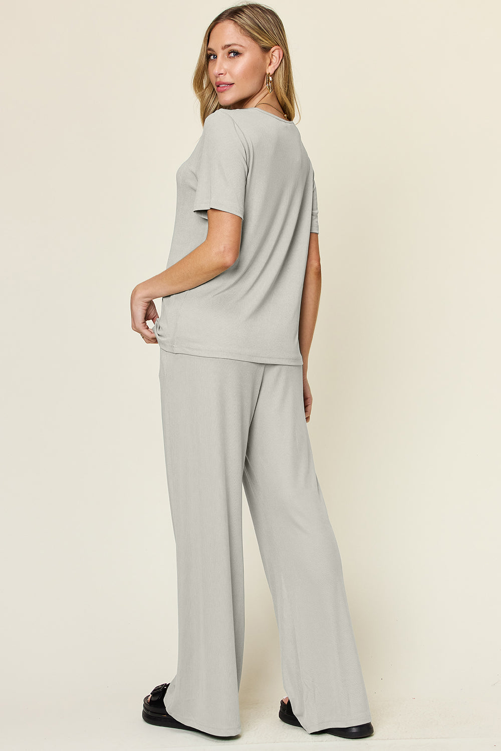 A person stands wearing the Double Take Full Size Round Neck Short Sleeve T-Shirt and Wide Leg Pants Set in a stretchy light grey fabric, with one hand in a pocket and black sandals.