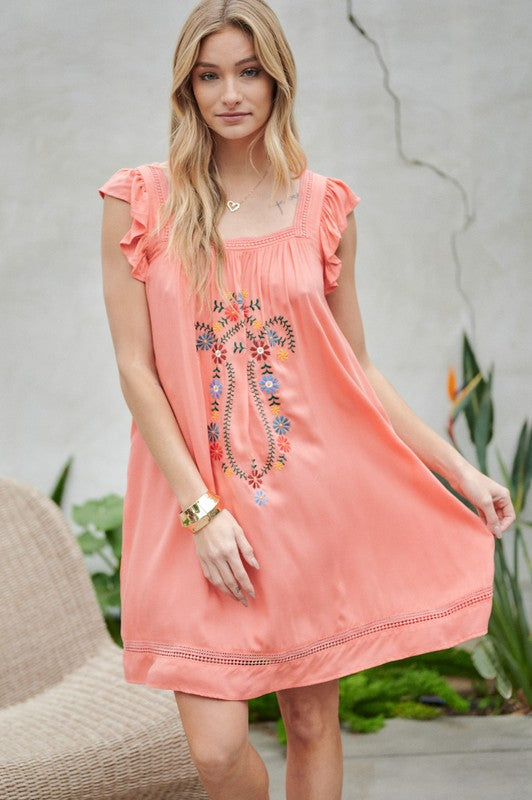 A woman in an Embroidered Square Neck Mini Dress, featuring a coral hue, stands near plants adjusting her hair—the perfect summer attire.