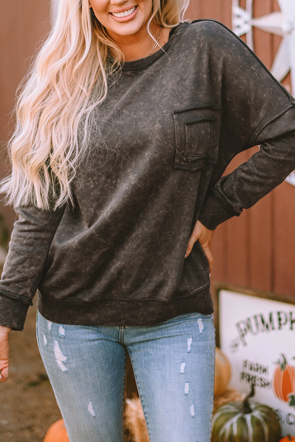 A person with long blonde hair stands with their back to the camera, wearing a Gray Acid Wash Drop Shoulder Long Sleeve Sweatshirt with Pockets and jeans. A large star decoration is visible in the background, along with some pumpkins on the ground.