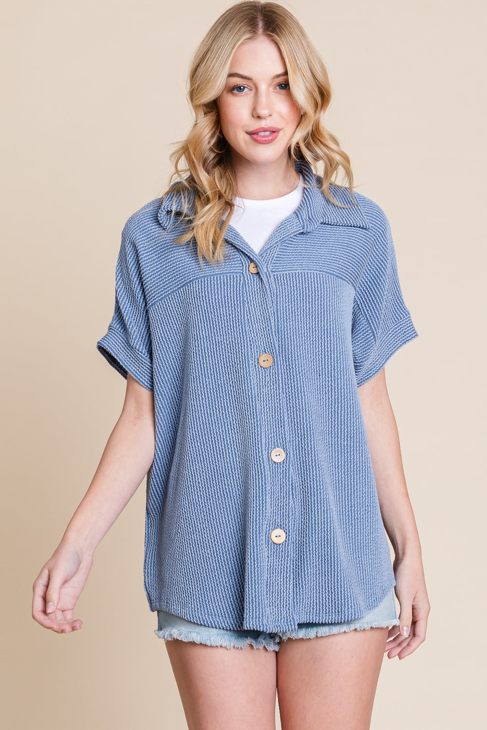 A woman wearing a BOMBOM Button Up Short Sleeves Ribbed Shirt in blue over a white shirt, paired with light denim shorts, stands against a beige background.