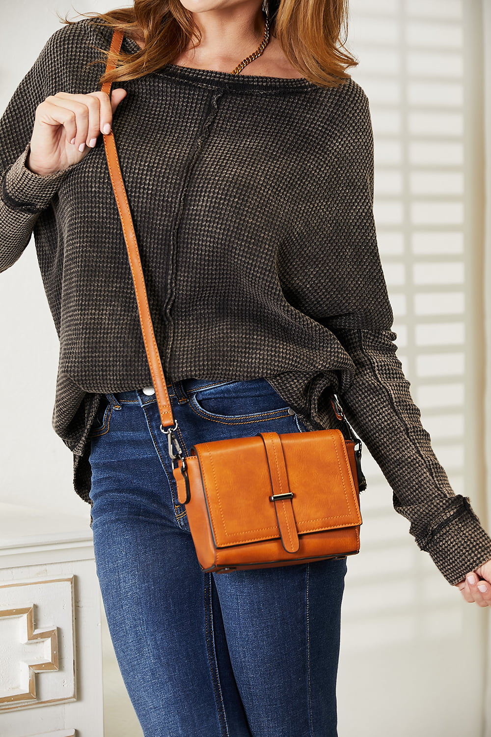 A person wearing a dark sweater and jeans holds the SHOMICO PU Leather Crossbody Bag, a stylish accessory in orange-brown featuring a flap and buckle detail.