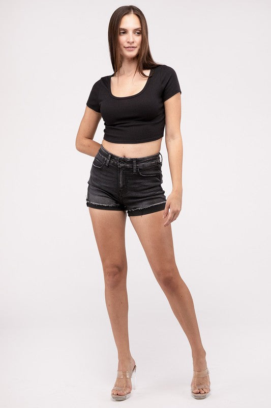 A person wearing washed black cuffed raw hem denim shorts stands with hands on hips against a neutral background.