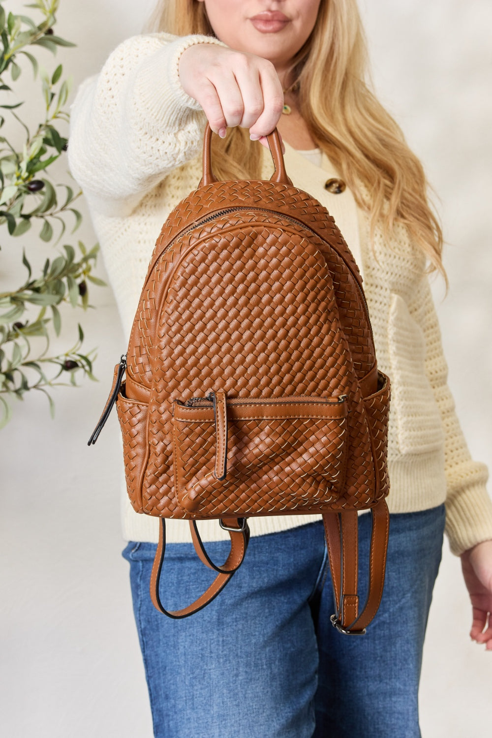 A person wearing a cream sweater and jeans is carrying the SHOMICO PU Leather Woven Backpack in brown.