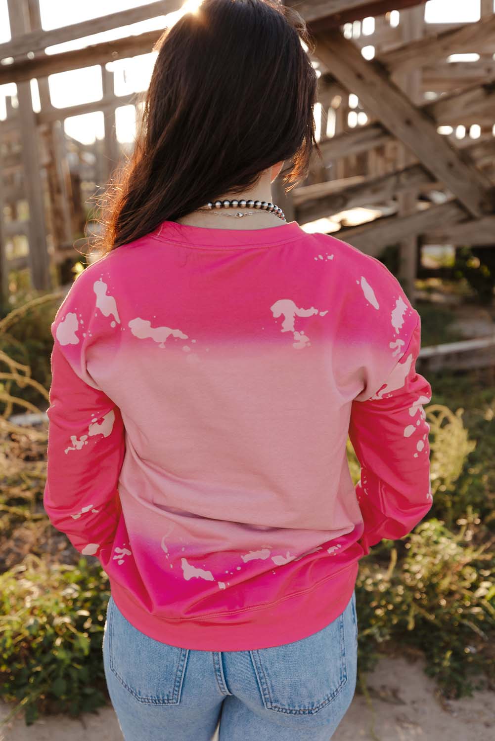 A person with long dark hair is standing outdoors, facing away from the camera, wearing a Pink Mama Rhinestone Letter Graphic Tie Dye Pullover and light blue jeans. Old wooden structures are visible in the background.