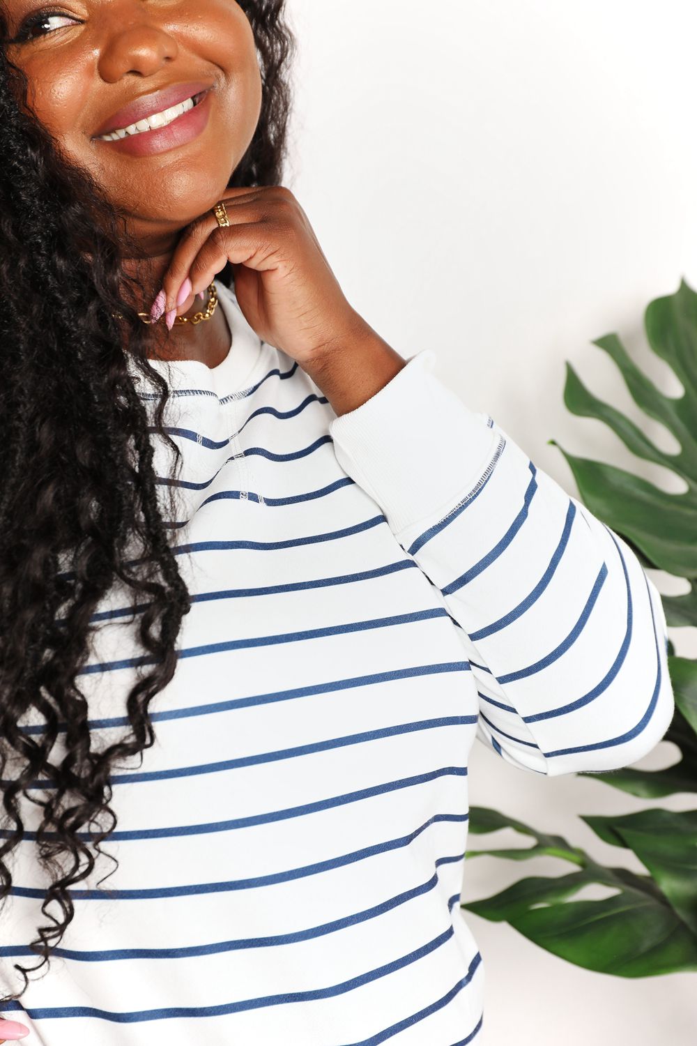 A person smiling while wearing the Double Take Striped Long Sleeve Round Neck Top, paired with denim shorts, stands in front of a white background with a plant.