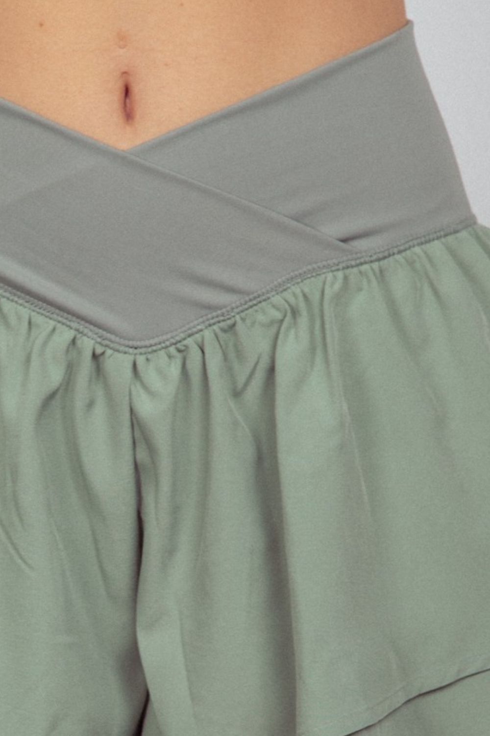 A close-up of a person wearing a light purple cropped top and sage green VERY J V-Shaped High Waist Layered Active Shorts with a crossover waistband.