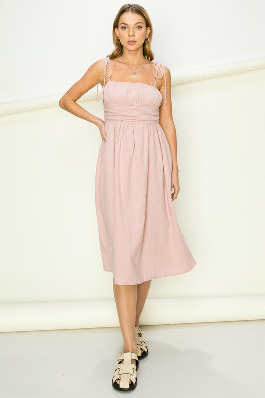 A woman wearing the Get a Clue Tie-Strap Midi Dress, featuring a light pink hue and tie-straps, showcases chic fashion and a flowy silhouette as she stands against a plain background.