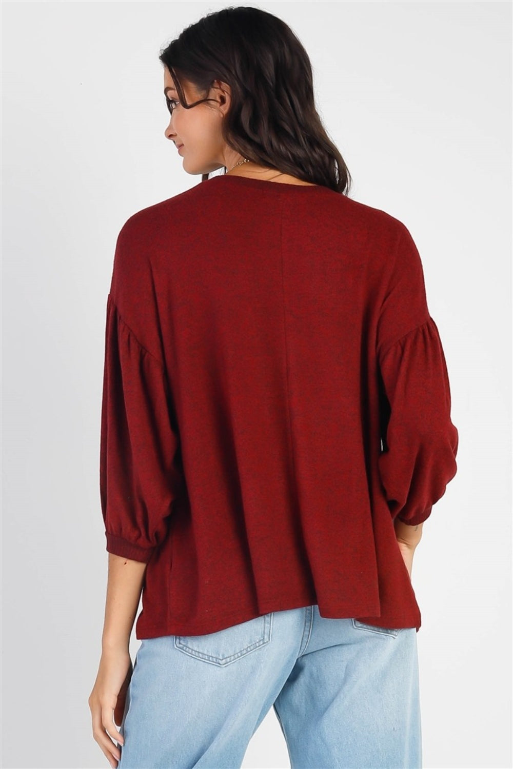 A person with long dark hair stands against a white background, smiling and raising one hand while wearing the trendy and stylish Cherish Apparel Drop Shoulder Puff Sleeve Top in burgundy paired with light blue jeans.