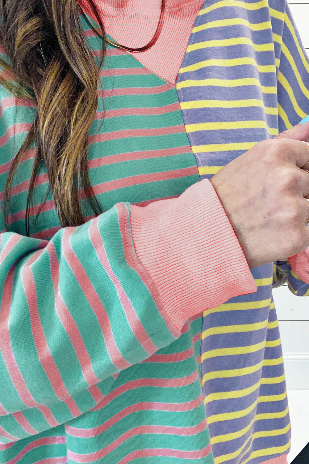 A person with wavy hair is seen from behind wearing a Casual Stripe Colorblock Drop Shoulder Oversize Sweatshirt in pastel colors and blue jeans. They are carrying a white shoulder bag, showcasing a trendy color block design.