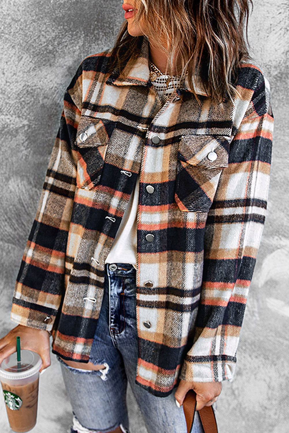 A person wearing a Brown Plaid Button Up Long Sleeve Flannel Shacket layered over a white top with ripped jeans and holding a brown handbag stands against a textured gray wall.