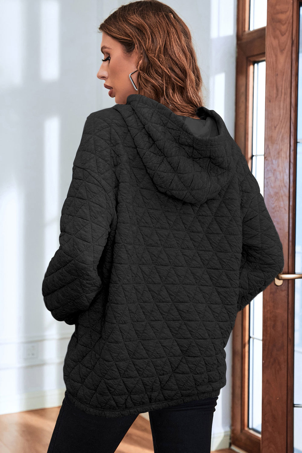 A person with wavy brown hair is standing indoors, facing a window, and wearing a Black Solid Color Quilted Kangaroo Pocket Hoodie with a drop shoulder design.